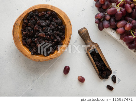 Sweet dark raisins in wooden bowl and scoop with red grapes in wooden box on light kitchen background. Sweet dark raisins in wooden bowl and scoop with red grapes in wooden box on light kitchen background. 115987471