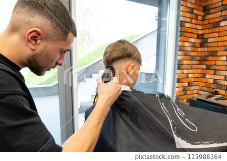 The picture of a blonde child with blue eyes and freckles is in a barbershop beauty salon. The miller holds a milling trimmer in his hand. The picture of a blonde child with blue eyes and freckles is in a barbershop beauty salon. The miller holds a milling trimmer in his hand. 115988164
