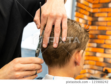 Man barber cutting little boy's hair using comb and scissors. Child getting haircut from adult male, likely barber. Professional hairdresser and cute client at modern barbershop. Man barber cutting little boy's hair using comb and scissors. Child getting haircut from adult male, likely barber. Professional hairdresser and cute client at modern barbershop. 115988242