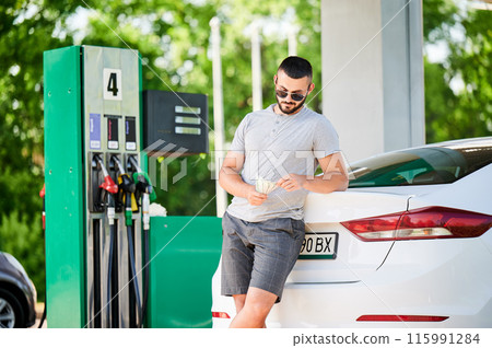 Young man waiting for petrol station operator to pay for fuel. Man leaning on his car and counting how much gasoline costs. Male in casual clothes holding money. Young man waiting for petrol station operator to pay for fuel. Man leaning on his car and counting how much gasoline costs. Male in casual clothes holding money. 115991284