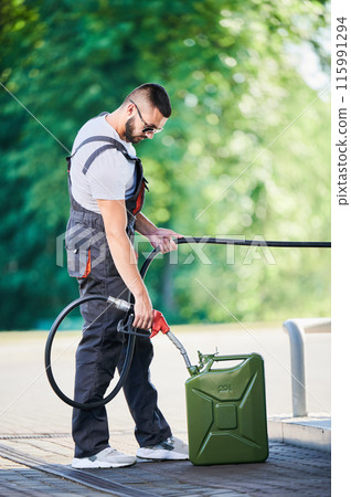 Professional worker refueling canister with fuel. Skilled man filling cistern with gasoline for customer. Man in ovealls with pump nozzle refueling canister next to gasoline column. Professional worker refueling canister with fuel. Skilled man filling cistern with gasoline for customer. Man in ovealls with pump nozzle refueling canister next to gasoline column. 115991294