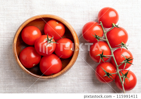 Red cherry tomatoes in a wooden bowl on linen fabric. Fresh, ripe type of small and round cocktail tomatoes, on the left side still on the vine, from above. Solanum lycopersicum var. cerasiforme. 115991411