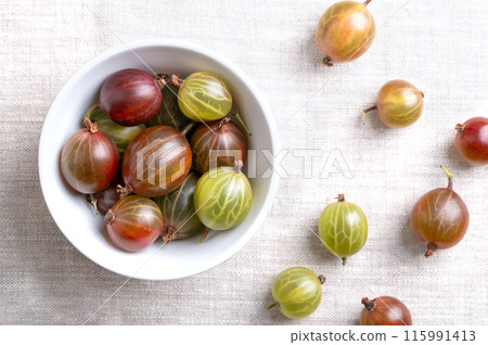 Green and red gooseberries in a white bowl on linen fabric. Fresh ripe berries, fruits of Ribes uva-crispa, known as European gooseberry, with sourish sweet taste. Close-up, from above, food photo. 115991413