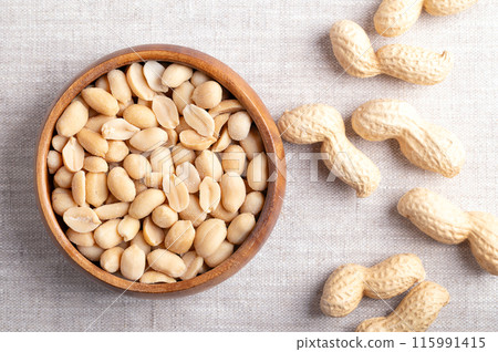 Roasted and salted peanuts in a wooden bowl on linen fabric. Ready-to-eat snack food, made from fruits of Arachis hypogaea, also known as groundnut, goober, pindar or monkey nut. Close-up from above. 115991415
