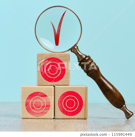 Wooden cubes with red targets stacked on top of each other. On the top cube there is a magnifying glass, through which you can see the red check mark 115991449