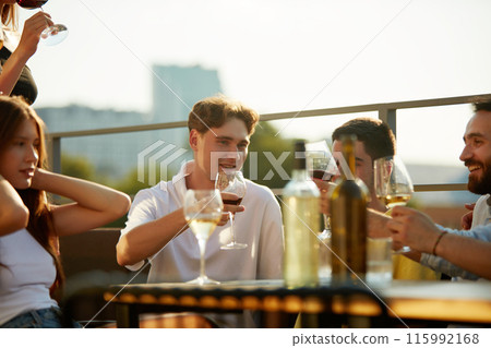 Sun casts golden glow on group of young people, friends as they raise their drinks in celebratory toast. 115992168