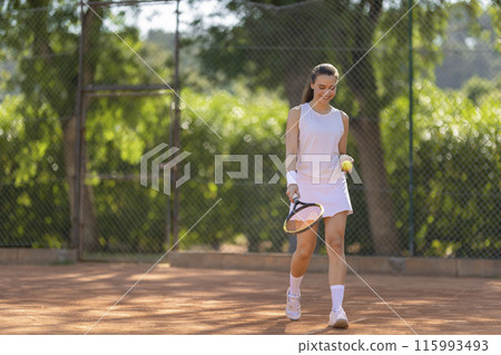 A woman is walking on a tennis court holding a tennis racket and a tennis ball 115993493