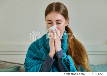 Young sick woman sitting under warm blanket sneezing coughing in handkerchief Young sick woman sitting under warm blanket sneezing coughing in handkerchief 115994223