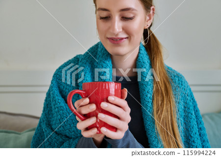 Young woman warming under blanket with mug of hot drink sitting on couch at home Young woman warming under blanket with mug of hot drink sitting on couch at home 115994224