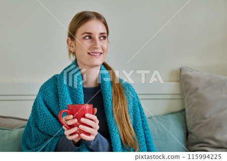Young woman warming under blanket with mug of hot drink sitting on couch at home 115994225
