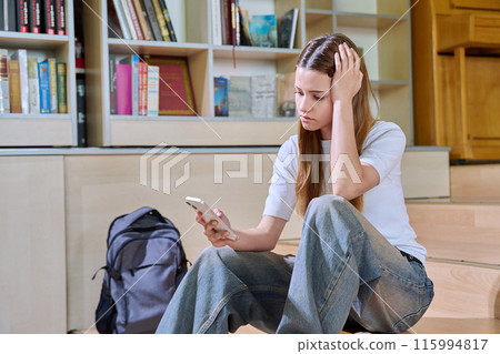 Sad teenage girl student looking at smartphone screen inside high school classroom 115994817