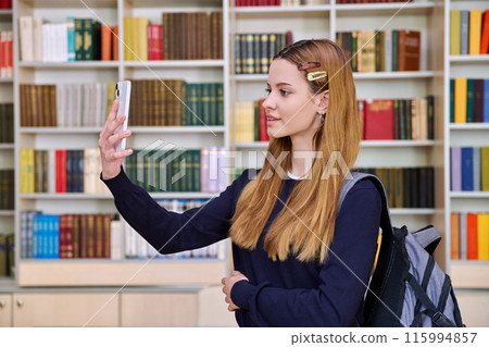 Teenage high school student using smartphone for video call chat, in library 115994857