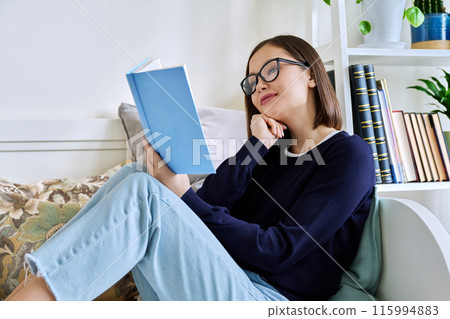 Young woman reading book, sitting on couch at home, engrossed in fiction 115994883