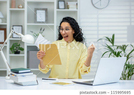 Happy woman celebrating after receiving good news in letter while working from home office. Expression of joy and success with laptop and books on the desk. Happy woman celebrating after receiving good news in letter while working from home office. Expression of joy and success with laptop and books on the desk. 115995695