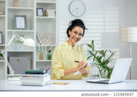 Woman student working from home, smiling and taking notes at a desk with a laptop. Bright and organized home office environment. 115995697