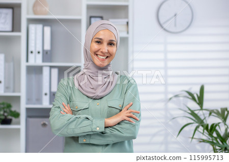 Smiling businesswoman in green shirt and headscarf standing in modern office with arms crossed. Professional confident pose with background of office shelves and clock Smiling businesswoman in green shirt and headscarf standing in modern office with arms crossed. Professional confident pose with background of office shelves and clock 115995713