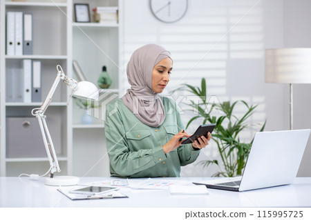 Female financial accountant calculates data with calculator while working at modern office desk with laptop and documents. Professional focusing on achieving accuracy in finances. Female financial accountant calculates data with calculator while working at modern office desk with laptop and documents. Professional focusing on achieving accuracy in finances. 115995725