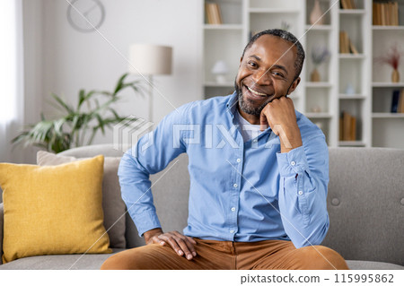 Smiling man sitting on couch in modern living room. Person wears blue shirt and brown pants, looking relaxed and happy. Bright and inviting home interior with plants, bookshelves, and decor Smiling man sitting on couch in modern living room. Person wears blue shirt and brown pants, looking relaxed and happy. Bright and inviting home interior with plants, bookshelves, and decor 115995862