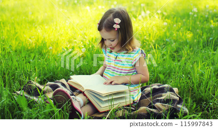 Happy little girl child reading book sitting on the grass in summer park Happy little girl child reading book sitting on the grass in summer park 115997843