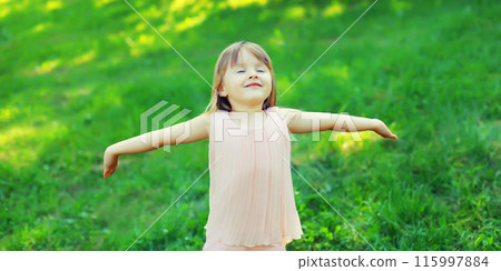 Happy little girl child enjoying warm weather standing on the grass in summer green park Happy little girl child enjoying warm weather standing on the grass in summer green park 115997884