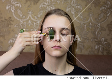 Young girl does eyelid massage using roller from natural stone to take care of skin and smooth out wrinkles, procedure for healing and caring for the skin of beautiful girl face 115999419