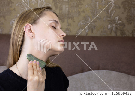 Young girl takes care of skin on her neck using special gua sha scraper for skin care, facial massage procedure for beautiful girl at home 115999432