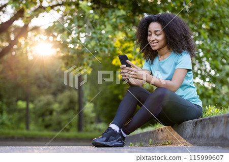 Smiling woman sitting on park curb after exercise, checking smartphone during sunset. Wearing activewear, enjoying outdoor workout, and surrounded by nature 115999607
