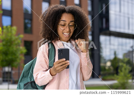 Young female student wearing glasses and backpack celebrating good news while looking at smartphone near university campus. Outdoor scene with modern building in background. Young female student wearing glasses and backpack celebrating good news while looking at smartphone near university campus. Outdoor scene with modern building in background. 115999621