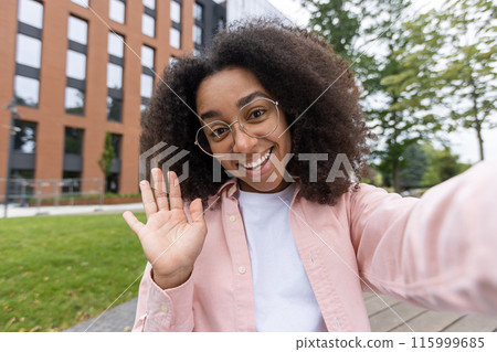 Smiling woman taking selfie outdoors on college campus. She is wearing a pink shirt and white t-shirt while waving at camera. Bright day with building and trees in background. Smiling woman taking selfie outdoors on college campus. She is wearing a pink shirt and white t-shirt while waving at camera. Bright day with building and trees in background. 115999685