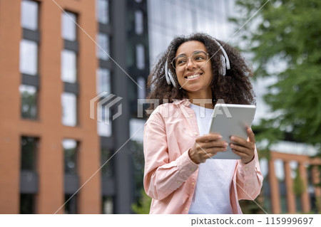 Happy young female student with headphones using tablet outside university campus. Student enjoying learning and music in modern educational environment. Happy young female student with headphones using tablet outside university campus. Student enjoying learning and music in modern educational environment. 115999697
