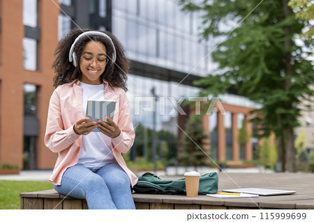 Young female student sitting near university campus using tablet with headphones on. Backpack, coffee cup, and notebooks on the table. Outdoor, casual setting with modern building in background. 115999699