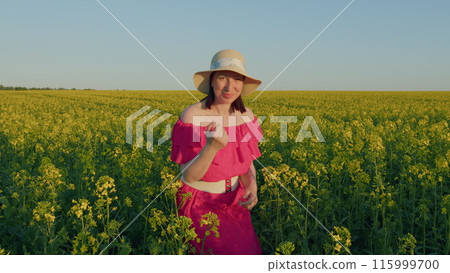 Pretty Woman Enjoying Blooming Rapeseed Field. Woman In Dress With Hat Walks In Green Field On Sunny Day. Romance Of Nature. Pretty Woman Enjoying Blooming Rapeseed Field. Woman In Dress With Hat Walks In Green Field On Sunny Day. Romance Of Nature. 115999700