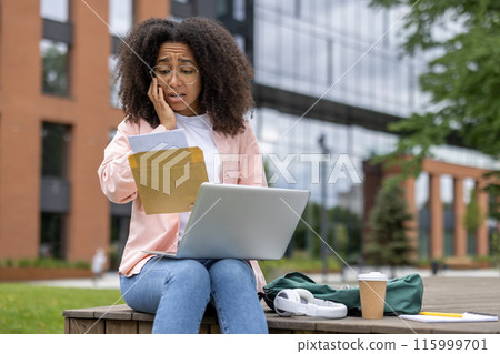 Young female student looking worried while reading letter with laptop outside university campus. Mixed emotions and stress evident in expression. 115999701