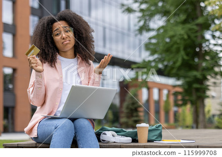 Young female student sitting near university campus with laptop and credit card, shrugging in confusion. Green glasses and pink shirt. Coffee cup, notebook, and backpack on bench. Young female student sitting near university campus with laptop and credit card, shrugging in confusion. Green glasses and pink shirt. Coffee cup, notebook, and backpack on bench. 115999708