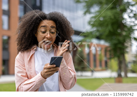 Young female student displaying excitement while looking at smartphone near university campus. Student with glasses and casual clothing expresses surprise and joy in outdoor setting. 115999742