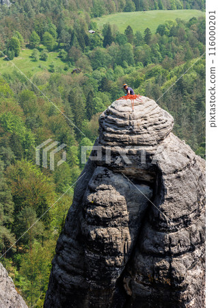 Tired rock climber resting break on peak summit rocky sansdtone mountain in Saxon Switzerland National Park Germany with green forest background. Extreme endurance sports adventure outdoors activity Tired rock climber resting break on peak summit rocky sansdtone mountain in Saxon Switzerland National Park Germany with green forest background. Extreme endurance sports adventure outdoors activity 116000201