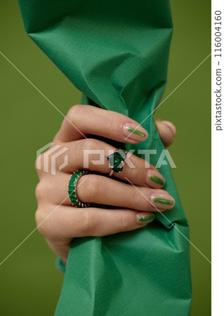 Vertical close up on composition of female hand with manicure of line design holding crumpled paper and wearing chic gemstone rings with emerald, on green background Vertical close up on composition of female hand with manicure of line design holding crumpled paper and wearing chic gemstone rings with emerald, on green background 116004160