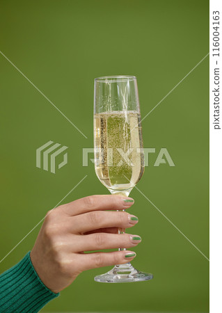 Vertical studio close up on female hand with nails of abstract design holding glass of champagne celebrating New Year or raising toast on fresh green background, copy space Vertical studio close up on female hand with nails of abstract design holding glass of champagne celebrating New Year or raising toast on fresh green background, copy space 116004163