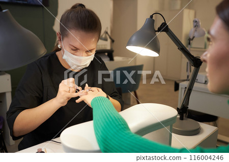 Shot of young female customer getting nails done and manicurist at work wearing face mask in beauty studio, copy space 116004246