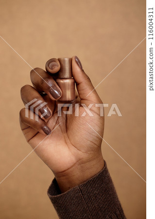 Vertical shot of Black woman holding golden nail polish for creating manicure. Dark brown nails pairing lacquer and skin tone of girl on beige background Vertical shot of Black woman holding golden nail polish for creating manicure. Dark brown nails pairing lacquer and skin tone of girl on beige background 116004311