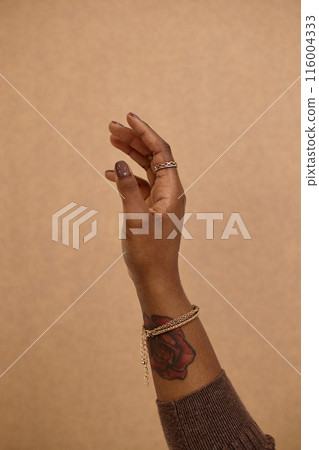 Vertical close up on female hand unrecognizable Black woman with dark brown nail and healthy skin, focus on tattooed female hand reaching up against neutral beige background 116004333