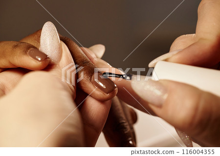 Close up shot of female nail master applying clear polish as last layer for manicure routine in salon. Final step transparent lacquer on top of dark brown polish coats, nail protection 116004355