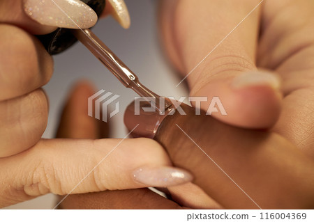 Macro shot of nail tech impeccable work using flat brush covering nail with dark brown lacquer. African American woman getting new nude nails in salon, copy space 116004369
