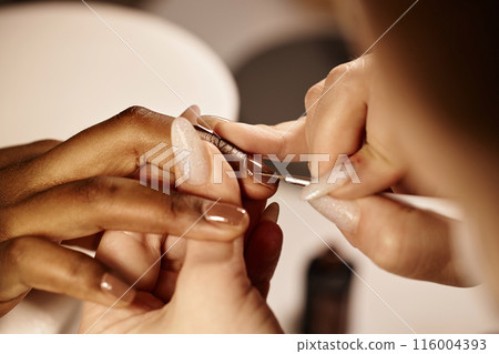 Close up on nail artists hands working on monochrome dark brown manicure using thin liner brush. African American woman getting nails done in beauty salon, copy space 116004393
