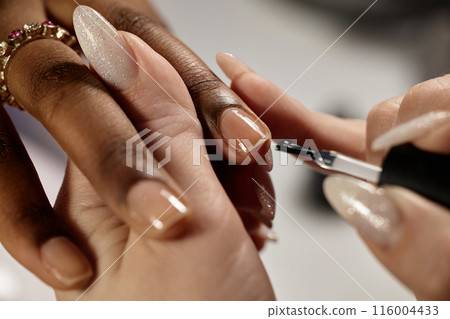 Close up on nail techs hands applying thin coat of clear nail polish on female clients fingernails in beauty salon 116004433