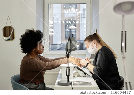 Side view shot of female nail master working on manicure of happy African American client with curly hair. Black woman at nail tech desk getting nails cut, filed, and shaped, copy space Side view shot of female nail master working on manicure of happy African American client with curly hair. Black woman at nail tech desk getting nails cut, filed, and shaped, copy space 116004457