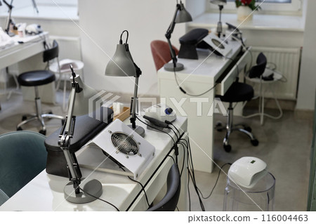 Shot of manicurist workplace equipped with nail tech desks with dust collectors and table lamps. White clean interior of empty beauty salon Shot of manicurist workplace equipped with nail tech desks with dust collectors and table lamps. White clean interior of empty beauty salon 116004463