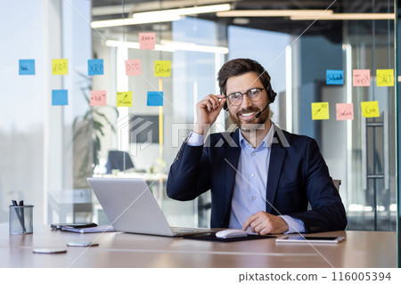Professional businessman wearing headset working on laptop in modern office with sticky notes on glass wall. Concept of remote work, customer service, and technology. 116005394