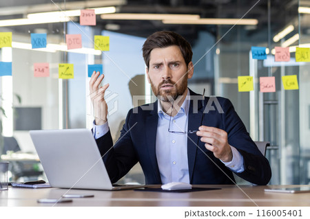 Frustrated businessman in office holding glasses, analyzing project on laptop, surrounded by colorful sticky notes. Expression of confusion, stress, and urgency in a professional setting 116005401