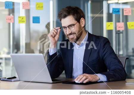 Smiling businessman working on laptop in modern office with sticky notes on glass wall. Professional in business attire focusing on work, planning, and strategy. Bright, collaborative workspace 116005408